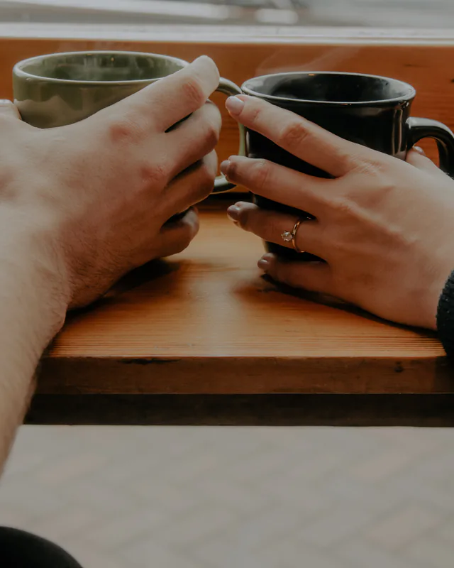 Dos personas con una taza de café en la mano, charlando en la barra de una cafetería; solo se ven las manos.